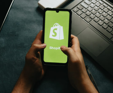 Hands holding a cell phone. On the screen of the cell phone is a neon green background with Shopify's white logo on top.