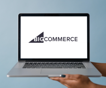 Two hands holding up the base of a mac laptop. On the screen fo the laptop is the logo for BigCommerce.